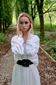 Young blonde woman in white skirt and shirt walks alone along the chestnut alley in the city park. Fashion woman. Young woman's modern portrait.