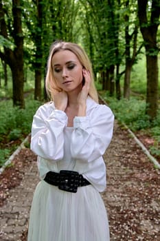 Young blonde woman in white skirt and shirt walks alone along the chestnut alley in the city park. Fashion woman. Young woman's modern portrait.