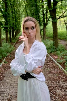 Young blonde woman in white skirt and shirt walks alone along the chestnut alley in the city park. Fashion woman. Young woman's modern portrait.