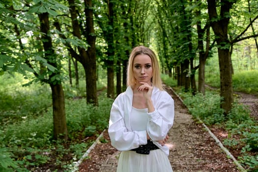 Young blonde woman in white skirt and shirt walks alone along the chestnut alley in the city park. Fashion woman. Young woman's modern portrait.