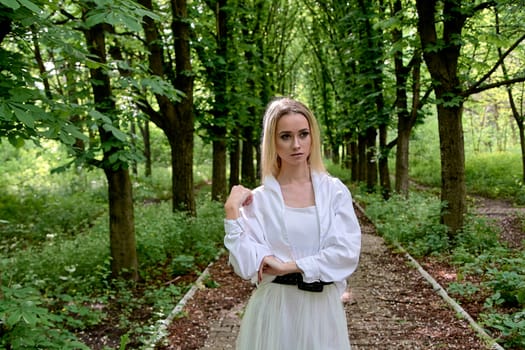 Young blonde woman in white skirt and shirt walks alone along the chestnut alley in the city park. Fashion woman. Young woman's modern portrait.