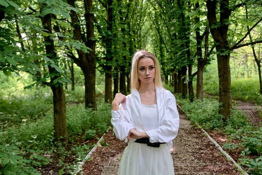 Young blonde woman in white skirt and shirt walks alone along the chestnut alley in the city park. Fashion woman. Young woman's modern portrait.