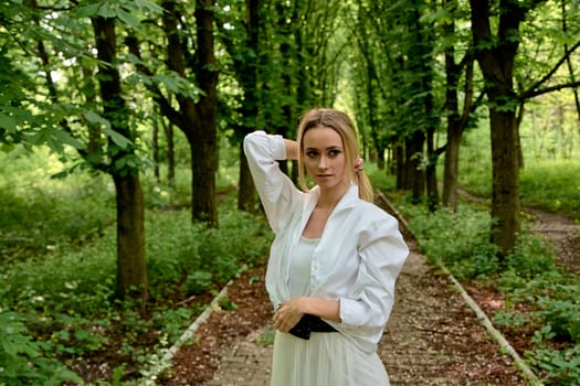 Young blonde woman in white skirt and shirt walks alone along the chestnut alley in the city park. Fashion woman. Young woman's modern portrait.