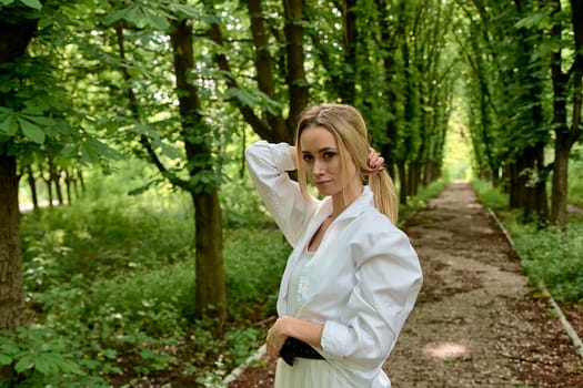 Young blonde woman in white skirt and shirt walks alone along the chestnut alley in the city park. Fashion woman. Young woman's modern portrait.