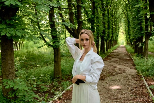 Young blonde woman in white skirt and shirt walks alone along the chestnut alley in the city park. Fashion woman. Young woman's modern portrait.