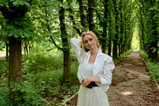 Young blonde woman in white skirt and shirt walks alone along the chestnut alley in the city park. Fashion woman. Young woman's modern portrait.