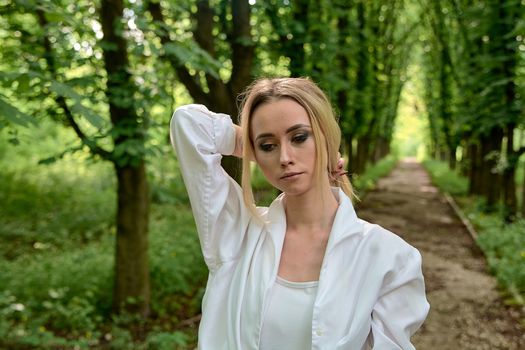 Young blonde woman in white skirt and shirt walks alone along the chestnut alley in the city park. Fashion woman. Young woman's modern portrait.