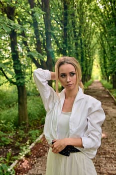 Young blonde woman in white skirt and shirt walks alone along the chestnut alley in the city park. Fashion woman. Young woman's modern portrait.