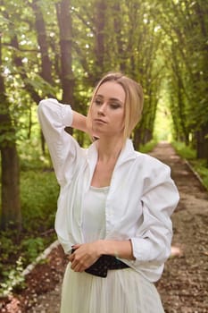 Young blonde woman in white skirt and shirt walks alone along the chestnut alley in the city park. Fashion woman. Young woman's modern portrait.