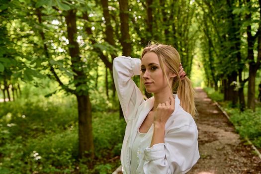 Young blonde woman in white skirt and shirt walks alone along the chestnut alley in the city park. Fashion woman. Young woman's modern portrait.