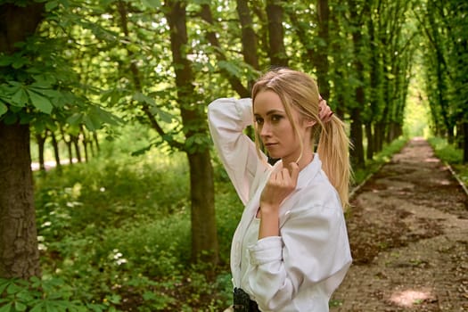 Young blonde woman in white skirt and shirt walks alone along the chestnut alley in the city park. Fashion woman. Young woman's modern portrait.