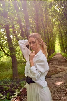 Young blonde woman in white skirt and shirt walks alone along the chestnut alley in the city park. Fashion woman. Young woman's modern portrait.