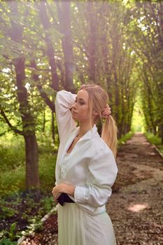 Young blonde woman in white skirt and shirt walks alone along the chestnut alley in the city park. Fashion woman. Young woman's modern portrait.