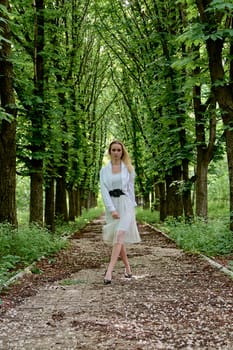 Young blonde woman in white skirt and shirt walks alone along the chestnut alley in the city park. Fashion woman. Young woman's modern portrait.