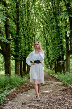 Young blonde woman in white skirt and shirt walks alone along the chestnut alley in the city park. Fashion woman. Young woman's modern portrait.