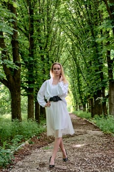 Young blonde woman in white skirt and shirt walks alone along the chestnut alley in the city park. Fashion woman. Young woman's modern portrait.