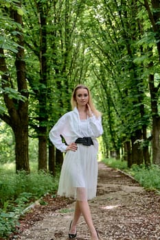 Young blonde woman in white skirt and shirt walks alone along the chestnut alley in the city park. Fashion woman. Young woman's modern portrait.