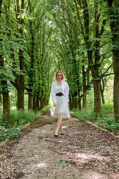 Young blonde woman in white skirt and shirt walks alone along the chestnut alley in the city park. Fashion woman. Young woman's modern portrait.