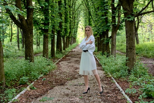 Young blonde woman in white skirt and shirt walks alone along the chestnut alley in the city park. Fashion woman. Young woman's modern portrait.