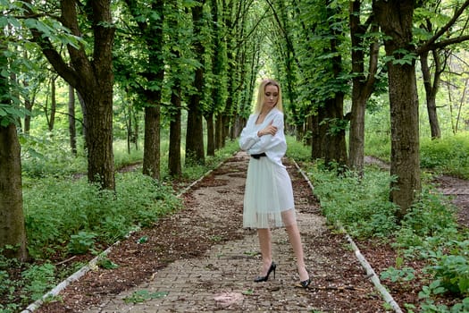 Young blonde woman in white skirt and shirt walks alone along the chestnut alley in the city park. Fashion woman. Young woman's modern portrait.