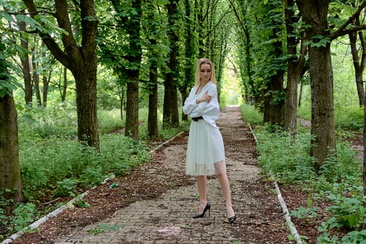 Young blonde woman in white skirt and shirt walks alone along the chestnut alley in the city park. Fashion woman. Young woman's modern portrait.