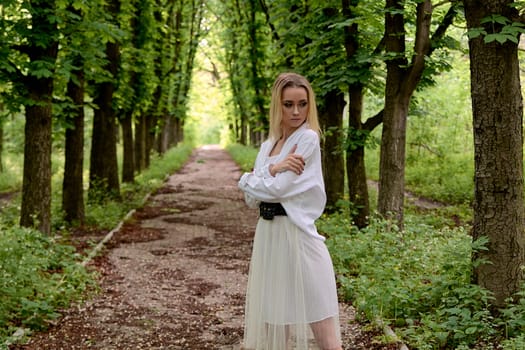 Young blonde woman in white skirt and shirt walks alone along the chestnut alley in the city park. Fashion woman. Young woman's modern portrait.