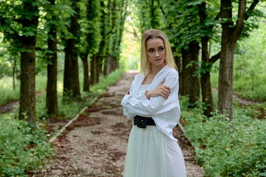 Young blonde woman in white skirt and shirt walks alone along the chestnut alley in the city park. Fashion woman. Young woman's modern portrait.