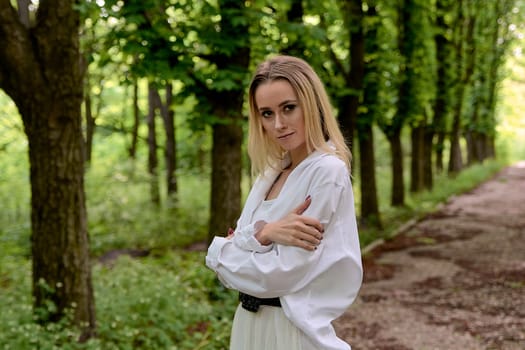 Young blonde woman in white skirt and shirt walks alone along the chestnut alley in the city park. Fashion woman. Young woman's modern portrait.