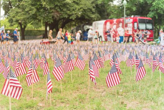 Close-up view of lawn American flag with row of people from Memorial Day March event in Dallas, Texas, USA. Blurry crowded family members carry fallen heroes banners pictures placards in parade