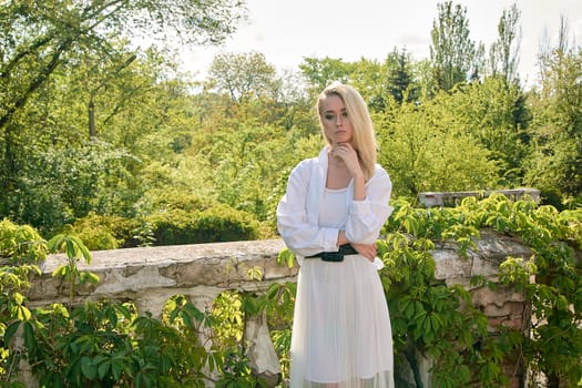 Young blonde woman in white skirt and shirt in the old sunny summer city park. Fashion woman. Young woman's modern portrait.