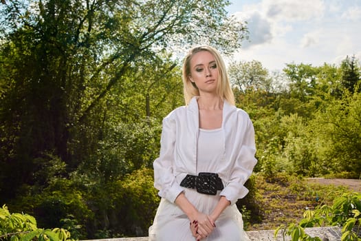 Young blonde woman in white skirt and shirt in the old sunny summer city park. Fashion woman. Young woman's modern portrait.