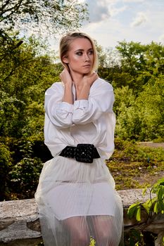 Young blonde woman in white skirt and shirt in the old sunny summer city park. Fashion woman. Young woman's modern portrait.