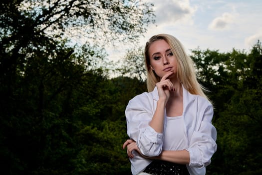 Young blonde woman in white skirt and shirt in the old sunny summer city park. Fashion woman. Young woman's modern portrait.