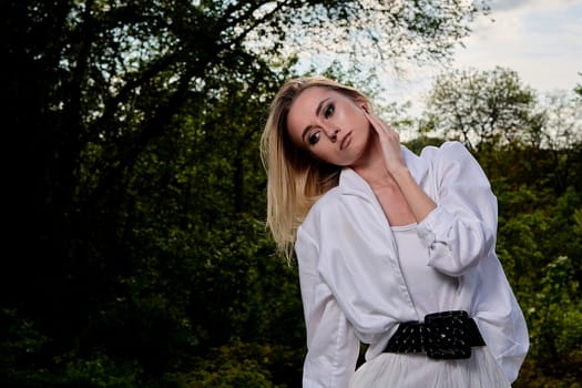 Young blonde woman in white skirt and shirt in the old sunny summer city park. Fashion woman. Young woman's modern portrait.