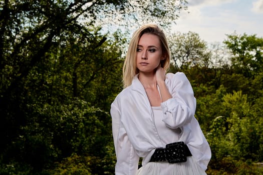 Young blonde woman in white skirt and shirt in the old sunny summer city park. Fashion woman. Young woman's modern portrait.