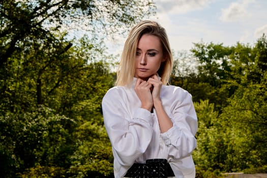 Young blonde woman in white skirt and shirt in the old sunny summer city park. Fashion woman. Young woman's modern portrait.