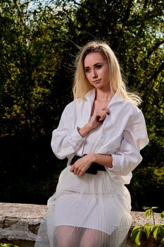 Young blonde woman in white skirt and shirt in the old sunny summer city park. Fashion woman. Young woman's modern portrait.