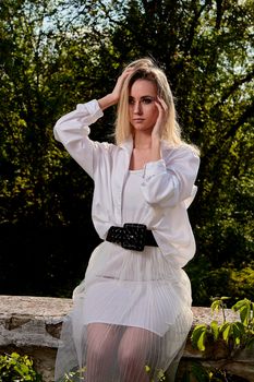 Young blonde woman in white skirt and shirt in the old sunny summer city park. Fashion woman. Young woman's modern portrait.