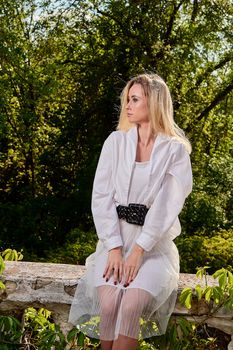 Young blonde woman in white skirt and shirt in the old sunny summer city park. Fashion woman. Young woman's modern portrait.