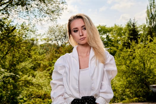 Young blonde woman in white skirt and shirt in the old sunny summer city park. Fashion woman. Young woman's modern portrait.