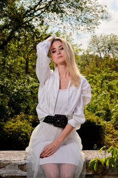 Young blonde woman in white skirt and shirt in the old sunny summer city park. Fashion woman. Young woman's modern portrait.