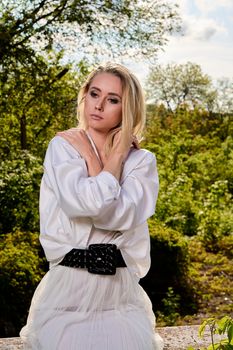 Young blonde woman in white skirt and shirt in the old sunny summer city park. Fashion woman. Young woman's modern portrait.