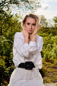 Young blonde woman in white skirt and shirt in the old sunny summer city park. Fashion woman. Young woman's modern portrait.