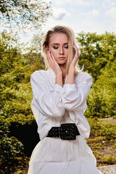 Young blonde woman in white skirt and shirt in the old sunny summer city park. Fashion woman. Young woman's modern portrait.