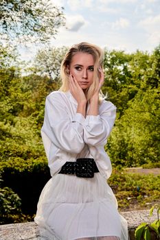 Young blonde woman in white skirt and shirt in the old sunny summer city park. Fashion woman. Young woman's modern portrait.