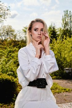 Young blonde woman in white skirt and shirt in the old sunny summer city park. Fashion woman. Young woman's modern portrait.