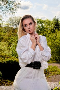 Young blonde woman in white skirt and shirt in the old sunny summer city park. Fashion woman. Young woman's modern portrait.