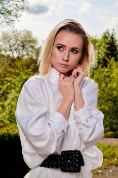 Young blonde woman in white skirt and shirt in the old sunny summer city park. Fashion woman. Young woman's modern portrait.