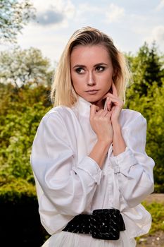 Young blonde woman in white skirt and shirt in the old sunny summer city park. Fashion woman. Young woman's modern portrait.