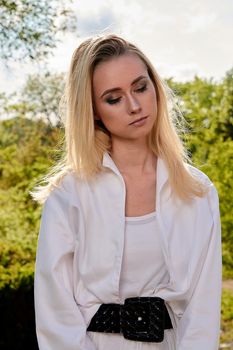 Young blonde woman in white skirt and shirt in the old sunny summer city park. Fashion woman. Young woman's modern portrait.